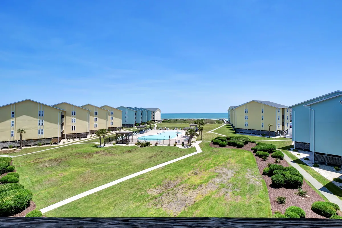 A vibrant exterior shot showcasing a sparkling swimming pool framed by landscaped gardens. The pool area overlooks the ocean, with nearby buildings providing context for the gated community. The scene exudes relaxation, with space for families to enjoy the sun or shade.