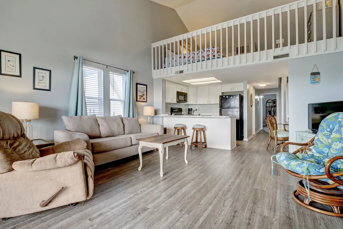 A cozy view of the living room with a sofa, kitchen area, and a visible loft bed overhead. The arrangement encourages family members or friends to engage in shared activities like cooking, relaxing, and socializing in one unified space.