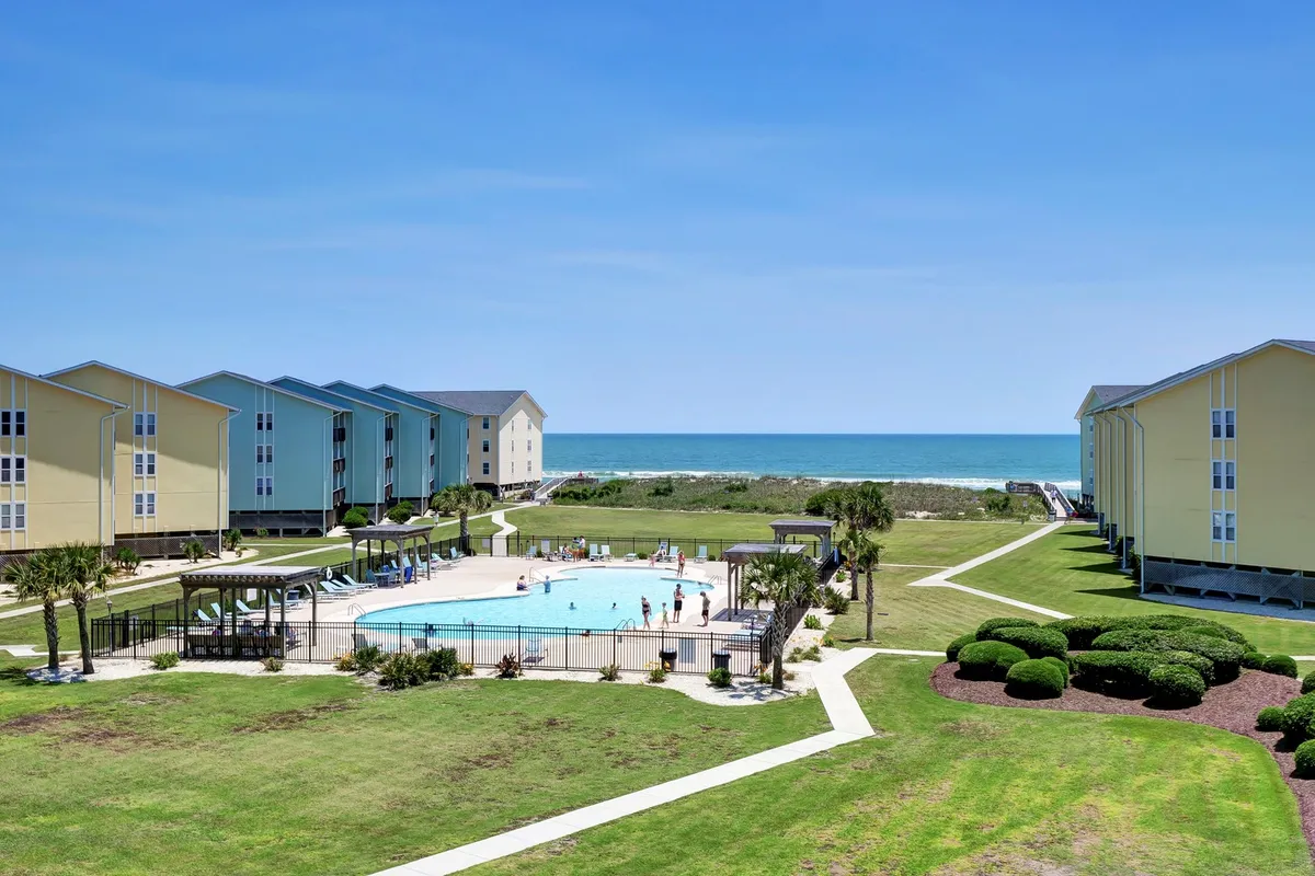 A wide view of the resort’s swimming pool framed by lush landscaped gardens, with the ocean visible just beyond. The scene includes the pool deck for lounging, glimpses of people sunbathing and socializing, and surrounding condo buildings that create a vibrant, communal atmosphere.