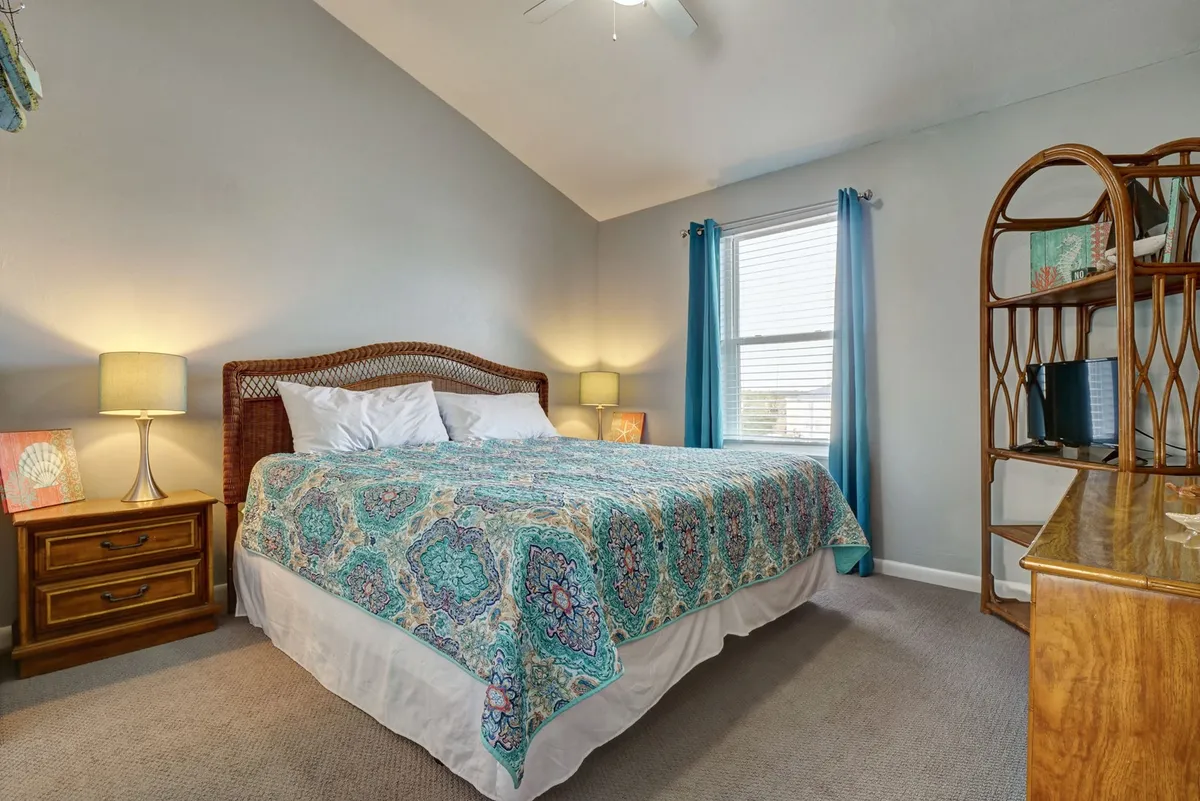 A bright, inviting bedroom featuring a queen-sized bed with colorful bedding, a rattan headboard, a nightstand with a lamp, a window dressed in light curtains, and a decorative bookshelf. The space feels cozy and serene, perfect for restful nights.
