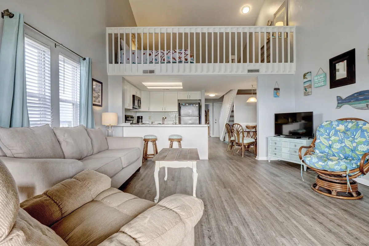This image captures a spacious, open-concept living area with a comfortable sofa, a lofted sleeping space perched above, a kitchen island with rattan chairs, and decorative wall art. The layout reveals how the sleeping, living, and kitchen areas blend seamlessly, emphasizing togetherness and communal living.