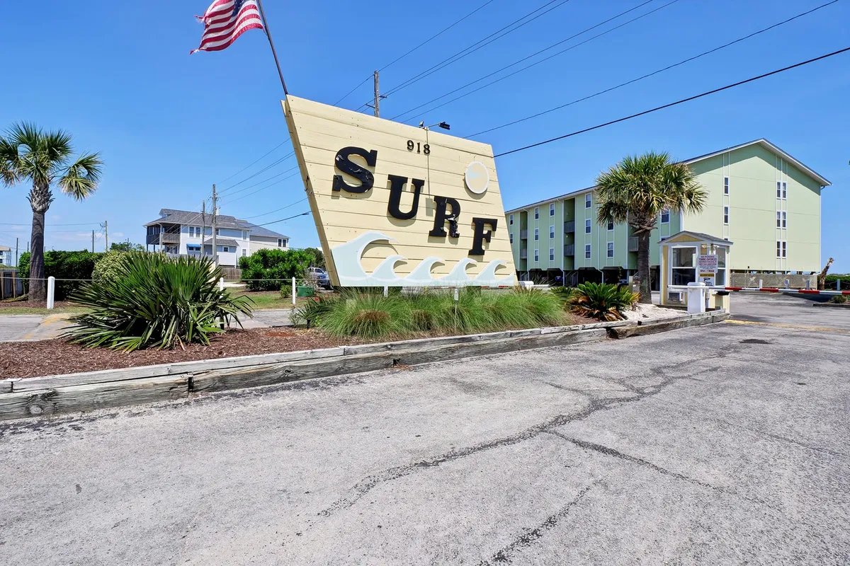 A cheerful exterior view featuring a large 'SURF' sign, tall palm trees, and multi-story buildings in the background. A flag waves in the breeze, and the atmosphere is lively, hinting at the surf-centric, welcoming community vibe and proximity to the beach.