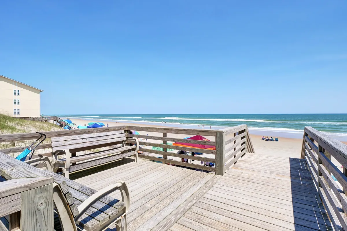 This exterior image features a spacious deck with comfortable seating overlooking the ocean, with beach umbrellas and sandy shoreline in view. The inviting setup suggests a place for guests to relax, sunbathe, or enjoy a coffee while taking in the coastal scenery. The vibrant ocean and open sky emphasize direct access to Surf City’s waterfront attractions.