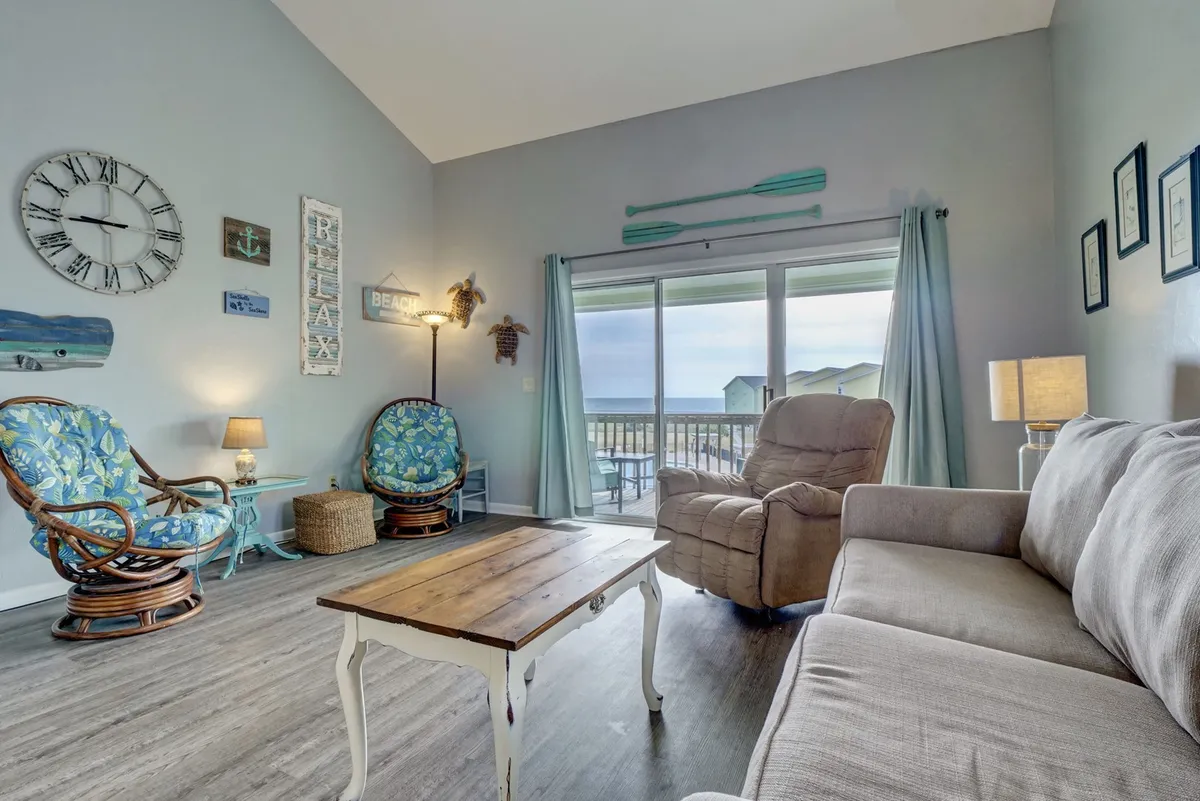 This inviting seating area features circular blue patterned chairs, a wooden coffee table, and a large window that offers expansive views—likely toward the courtyard, pool, or ocean. The space is designed for relaxed conversations, game nights, and enjoying scenic vistas from the third-floor vantage point.