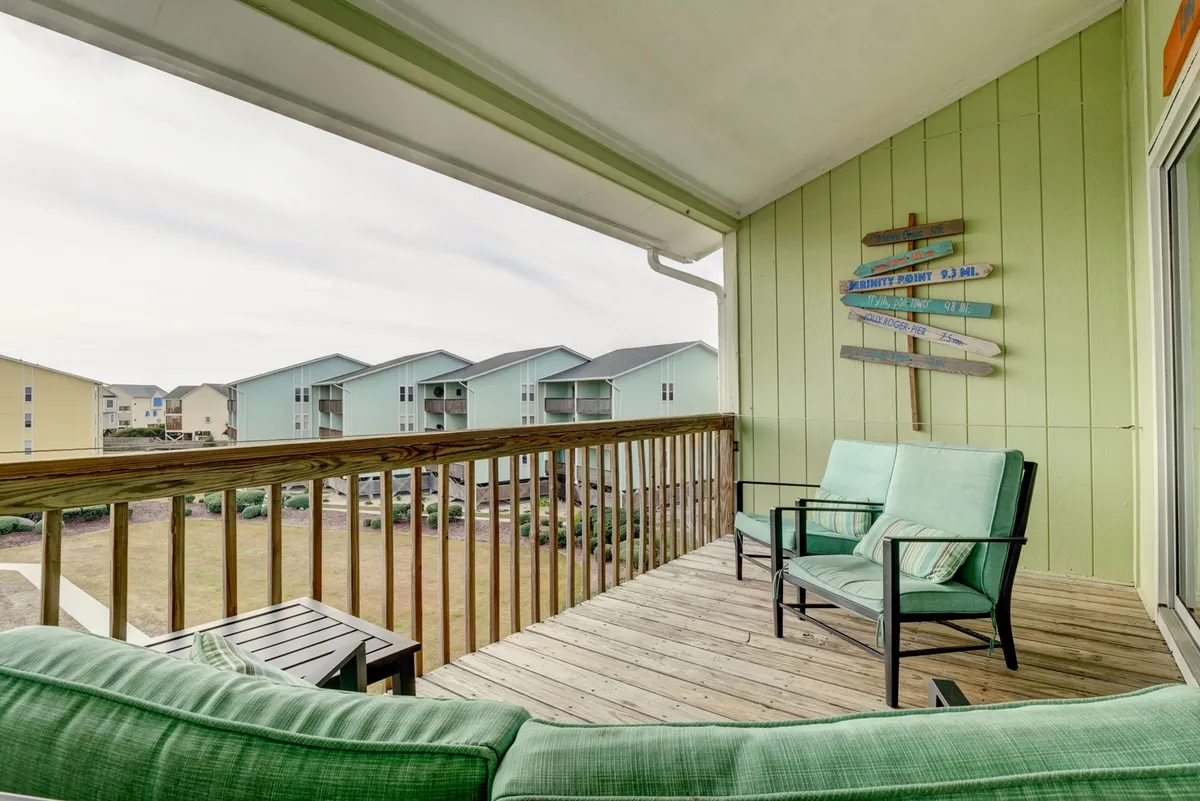 This exterior shot captures a green seating area with comfortable chairs arranged near a wooden railing, set against the backdrop of a tranquil outdoor environment. A directional sign hints at the community's resort-like feel, and the open-air layout suggests a perfect spot for socializing or unwinding while enjoying the fresh breeze.
