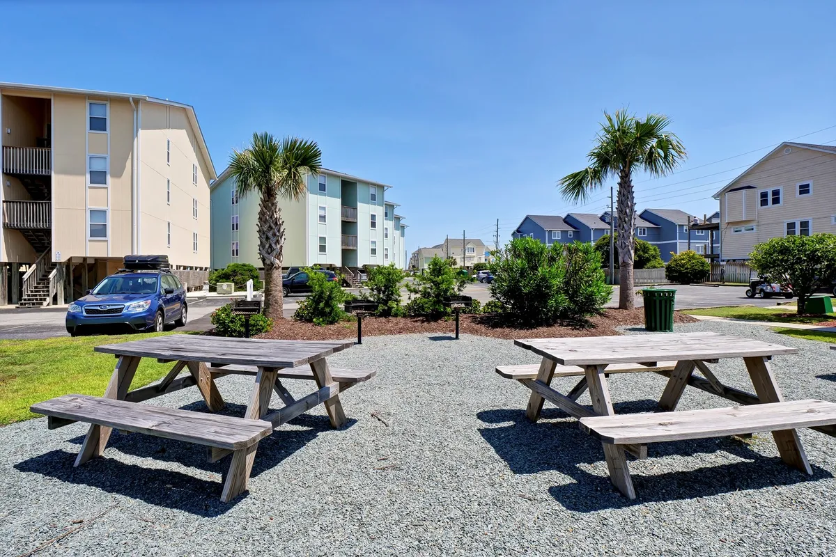 A shaded outdoor grilling and picnic area, framed by palm trees and surrounded by vibrant landscaping. Picnic tables invite guests to gather for meals or sunset socials, encapsulating the communal spirit and the joy of open-air dining after a day at the beach.