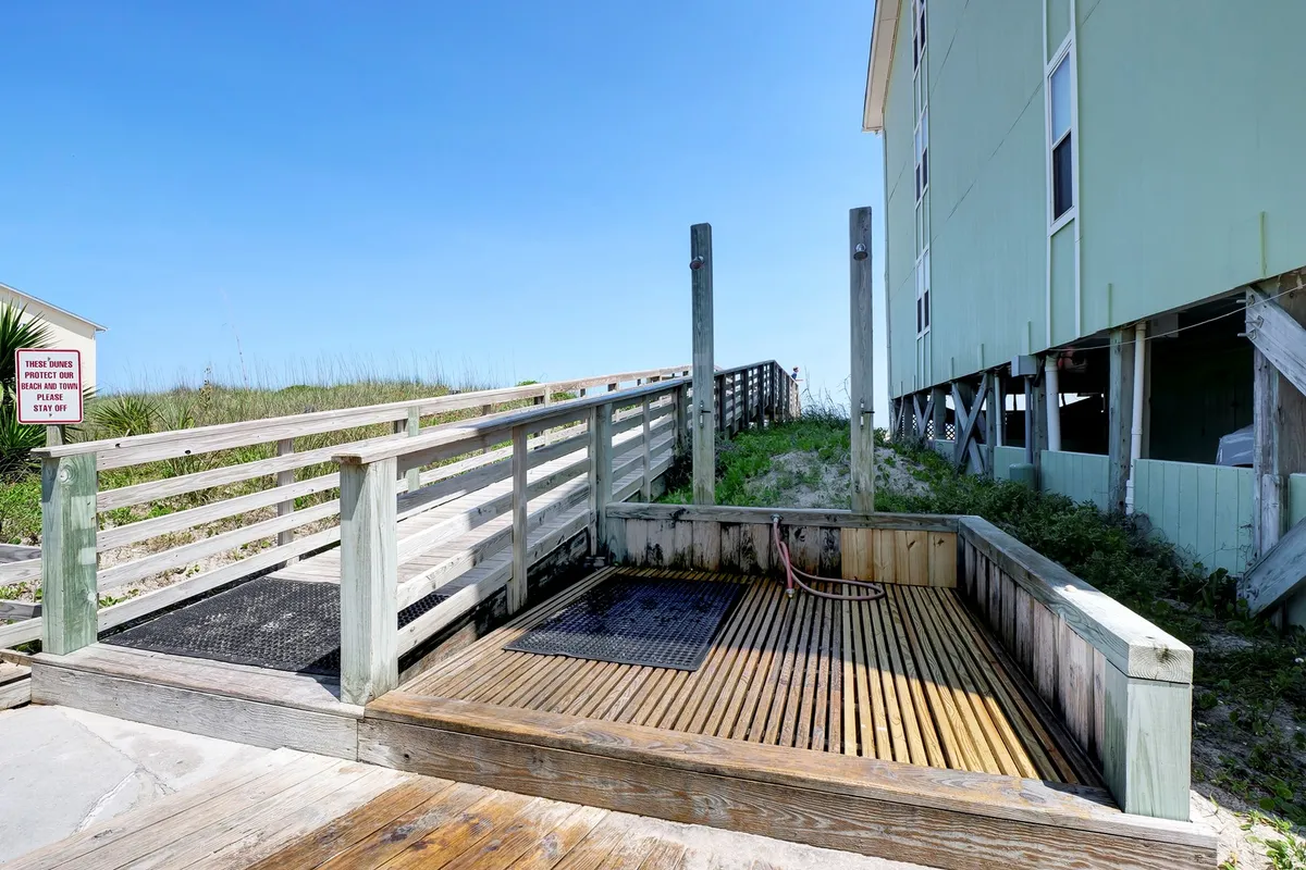 A wooden deck leading to a beach access ramp bordered by greenery, with a shower area for rinsing off after ocean swims. The deck’s vantage point highlights the ease of transitioning from the community to the sandy shore, and the relaxed gathering space families enjoy before and after beach adventures.