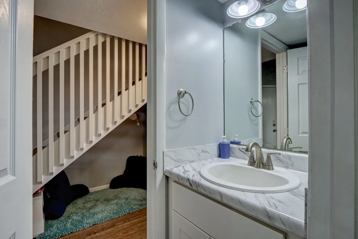 An interior view featuring a modern vanity sink with a marble countertop, a decorative mirror, and a staircase visible in the background. The area is well-lit, blending grooming space with the layout of the home.