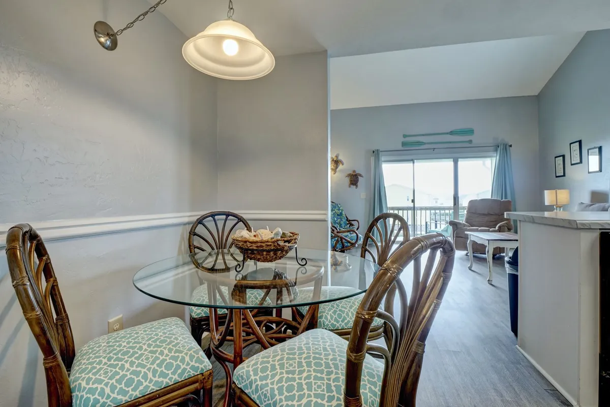 This image shows a bright, welcoming dining area with a glass table, woven basket centerpiece, blue accent cushions, and a large window that floods the space with natural light. The setup implies a casual, sociable atmosphere—perfect for shared meals or friendly gatherings after a day outdoors.